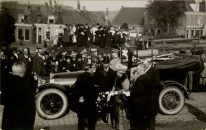 Zwart wit foto van koningin Wilhelmina, prins Hendrik en diverse hoogwaardigheidsbekleders bij een open auto met publiek op de achtergrond.